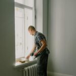 A man in overalls painting a window ledge inside a home, bright daylight.